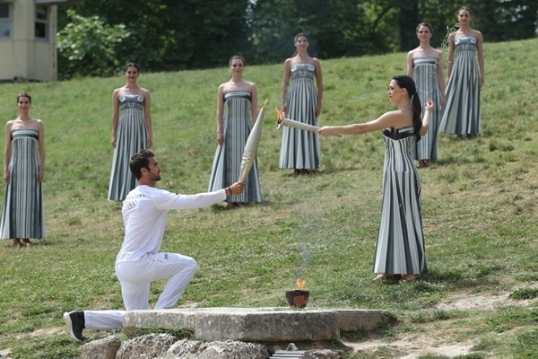 扮演最高女祭司的演員瑪麗·米娜（前右）點(diǎn)燃第一棒火炬手——希臘運(yùn)動(dòng)員、東京奧運(yùn)會(huì)男子賽艇項(xiàng)目金牌得主斯特凡諾斯·恩圖斯科斯手中的火炬。新華社記者 趙丁喆 攝